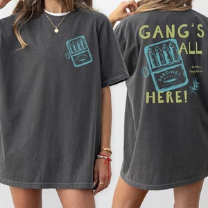 May include: A charcoal gray t-shirt featuring a small graphic of a sardine tin on the front left chest. The back displays a larger version of the same sardine tin graphic with the text "GANG'S ALL HERE!" in yellow and "better together" in white.