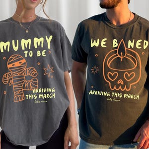 May include: Two dark gray t-shirts with matching designs. One says "MUMMY TO BE" with a mummy graphic, the other says "WE BANED" with a skull graphic. Both say "ARRIVING THIS MARCH".