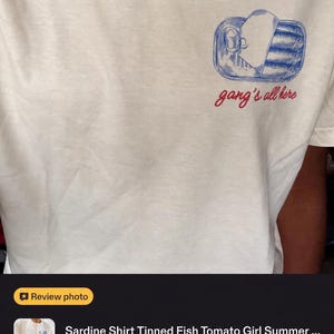May include: A cream-coloured t-shirt featuring a blue graphic of an open tin of sardines on the left chest. Below the illustration, the text "gang's all here" is printed in a red cursive font. The shirt has a crew neckline and short sleeves.