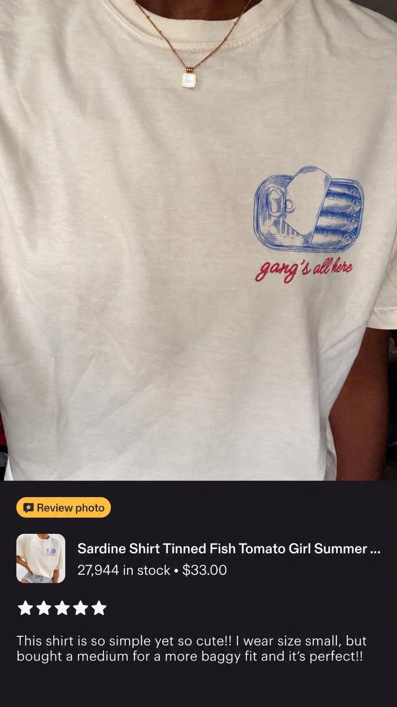 May include: A cream-coloured t-shirt featuring a blue graphic of an open tin of sardines on the left chest. Below the illustration, the text "gang's all here" is printed in a red cursive font. The shirt has a crew neckline and short sleeves.