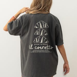 Op de afbeelding: Een donkergrijs T-shirt met een witte afbeelding van drie croissants die op elkaar gestapeld zijn. De tekst "BUON GIORNO il cornetto" is onder de croissants gedrukt. De tekst onder de afbeelding luidt "BEST ENJOYED WITH A PIPING HOT CAPPUCCINO MADE WITH ITALIAN ESPRESSO BEANS & FULL FAT MILK DISCLAIMER CAPPUCCINO SHOULD ONLY BE CONSUMED BEFORE 11AM"