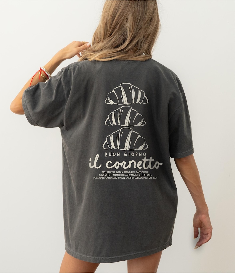 Op de afbeelding: Een donkergrijs T-shirt met een witte afbeelding van drie croissants die op elkaar gestapeld zijn. De tekst "BUON GIORNO il cornetto" is onder de croissants gedrukt. De tekst onder de afbeelding luidt "BEST ENJOYED WITH A PIPING HOT CAPPUCCINO MADE WITH ITALIAN ESPRESSO BEANS & FULL FAT MILK DISCLAIMER CAPPUCCINO SHOULD ONLY BE CONSUMED BEFORE 11AM"