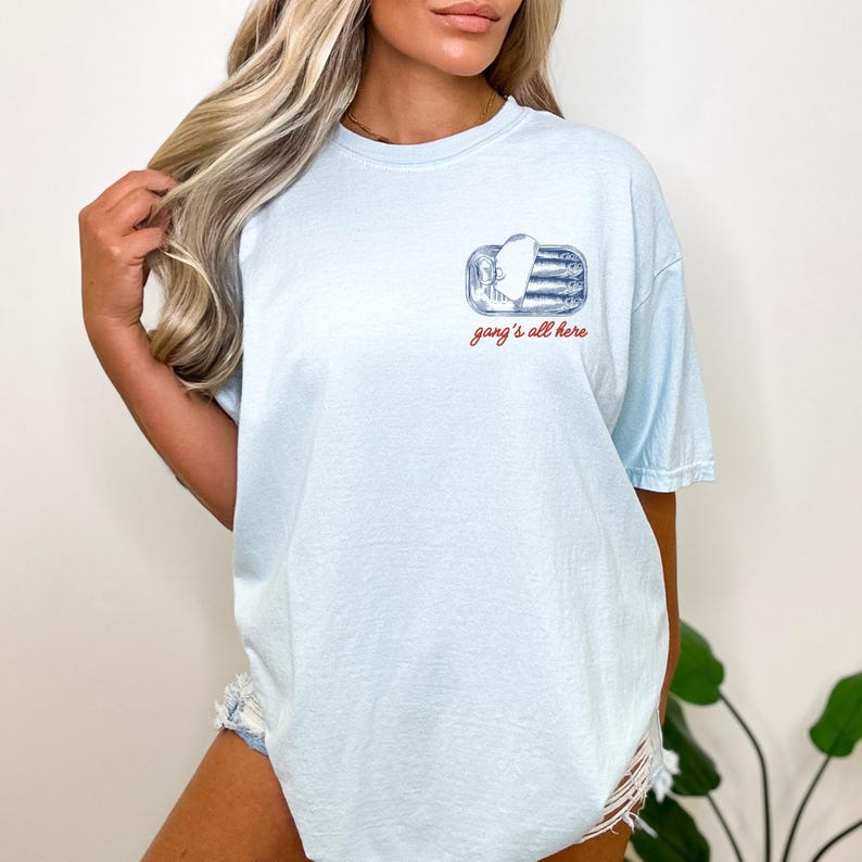 May include: A light blue short-sleeve t-shirt featuring a small graphic on the upper left chest. The graphic depicts a tin of sardines with the text "gang's all here" written in a cursive red font directly underneath the illustration.