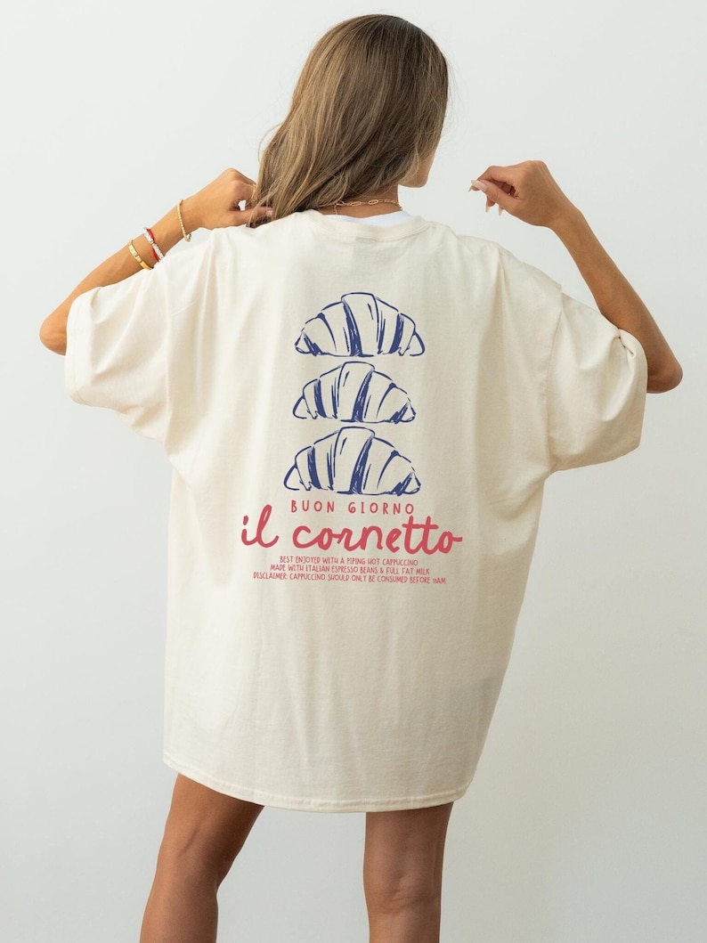 Op de afbeelding: Een cr&egrave;mekleurig T-shirt met een blauwe en rode afbeelding van croissants en de tekst "BUON GIORNO il cornetto"  BEST ENJOYED WITH A PIPING HOT CAPPUCCINO MADE WITH ITALIAN ESPRESSO BEANS & FULL FAT MILK DISCLAIMER: CAPPUCCINO SHOULD ONLY BE CONSUMED BEFORE HAM.