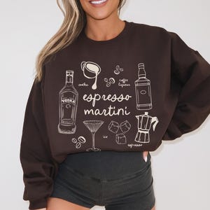 K&ouml;nnte beinhalten: Ein dunkelbraunes Sweatshirt mit einer wei&szlig;en Grafik, die ein Espresso-Martini-Rezept darstellt. Das Design umfasst Illustrationen von Wodka, Kaffeelik&ouml;r, Kaffeebohnen, Eisw&uuml;rfeln, einem Cocktailglas und einer Espressokanne. Die Worte "Espresso Martini" sind ebenfalls enthalten.