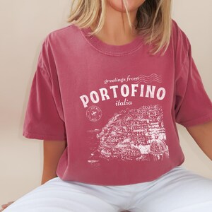 Może przedstawiać: Bordowy t-shirt z białym nadrukiem graficznym przedstawiającym panoramę miasta i tekstem "greetings from PORTOFINO italia".