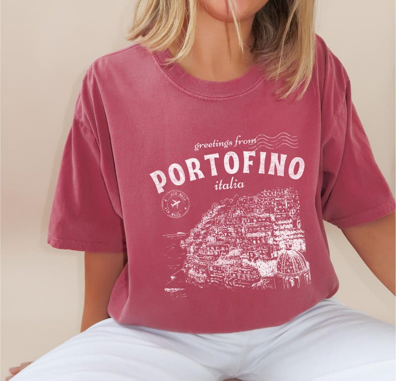 Może przedstawiać: Bordowy t-shirt z białym nadrukiem graficznym przedstawiającym panoramę miasta i tekstem "greetings from PORTOFINO italia".