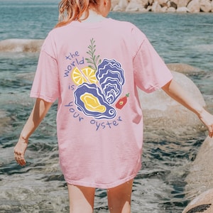 May include: A pink t-shirt with the text "the world is your oyster" and a graphic of an oyster, lemon, and hot sauce.