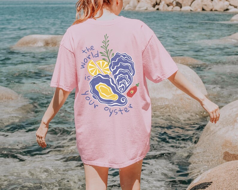 May include: A pink t-shirt with the text "the world is your oyster" and a graphic of an oyster, lemon, and hot sauce.