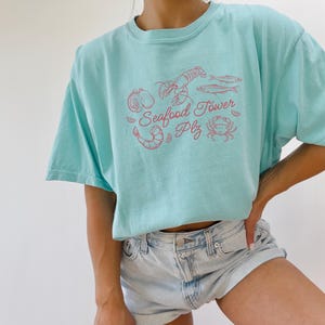 Peut inclure: Un t-shirt bleu clair avec un graphique rose de fruits de mer, y compris un homard, des crevettes, du poisson et un crabe. Le texte sur le t-shirt est "Seafood Tower Plz".