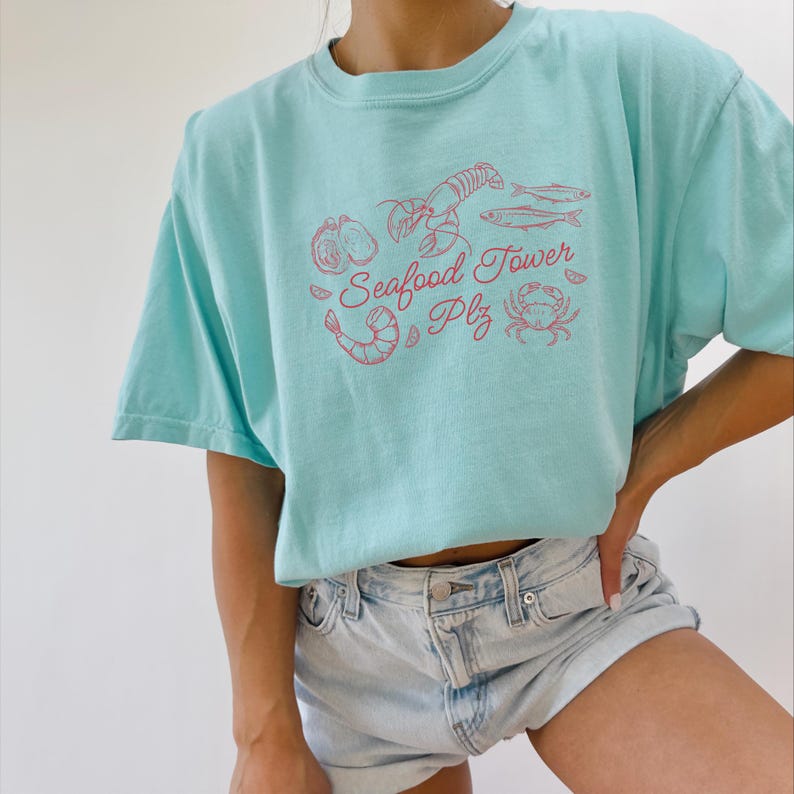 Peut inclure: Un t-shirt bleu clair avec un graphique rose de fruits de mer, y compris un homard, des crevettes, du poisson et un crabe. Le texte sur le t-shirt est "Seafood Tower Plz".