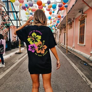 May include: Black t-shirt with a floral design and the text "LIFE IS BETTER AT THE BEACH" in neon colors. The shirt is worn by a person standing on a street with colorful paper lanterns overhead.