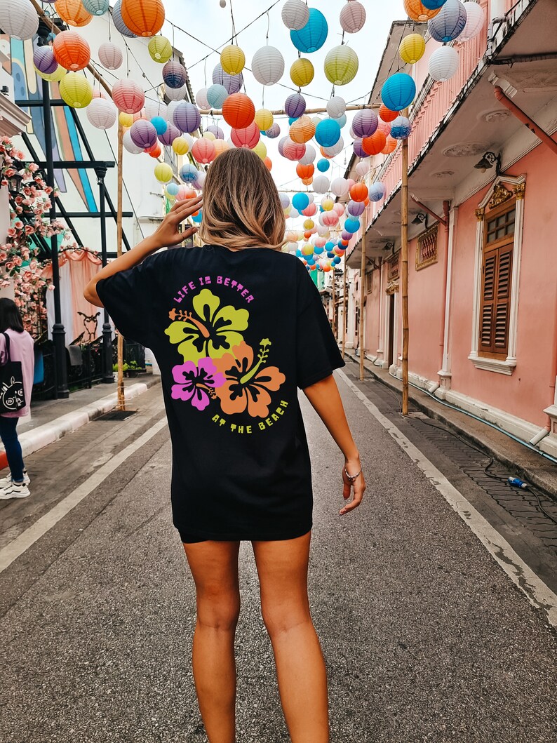 May include: Black t-shirt with a floral design and the text "LIFE IS BETTER AT THE BEACH" in neon colors. The shirt is worn by a person standing on a street with colorful paper lanterns overhead.