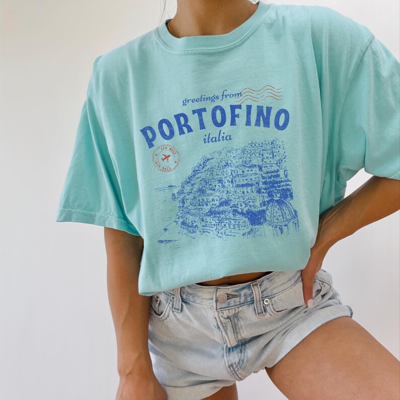 Może przedstawiać: Jasnoniebieska koszulka z nadrukiem graficznym przedstawiającym nadmorskie miasto z tekstem "greetings from PORTOFINO italia".