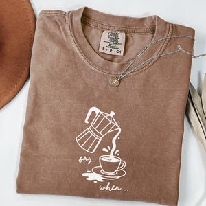 May include: A brown Comfort Colors t-shirt with a white graphic of a coffee pot pouring into a cup, with the words "say when..." below. A silver necklace with a small pendant is on the shirt.