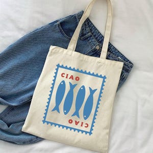 May include: A cream-colored tote bag with a blue and red graphic. The graphic features four blue fish inside a blue rectangular frame with the word "CIAO" in red above and below. The bag has two cream-colored handles and is placed on top of blue jeans.