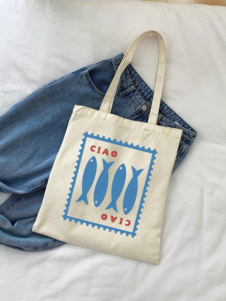 May include: A cream-colored tote bag with a blue and red graphic. The graphic features four blue fish inside a blue rectangular frame with the word "CIAO" in red above and below. The bag has two cream-colored handles and is placed on top of blue jeans.