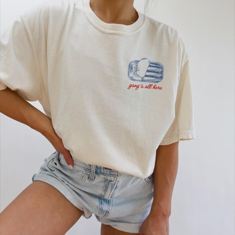 May include: Cream-colored t-shirt with a small graphic of a sardine can in blue and white, with the text "gang's all here" in red. The shirt has a relaxed fit and short sleeves. The model is wearing light blue denim shorts.