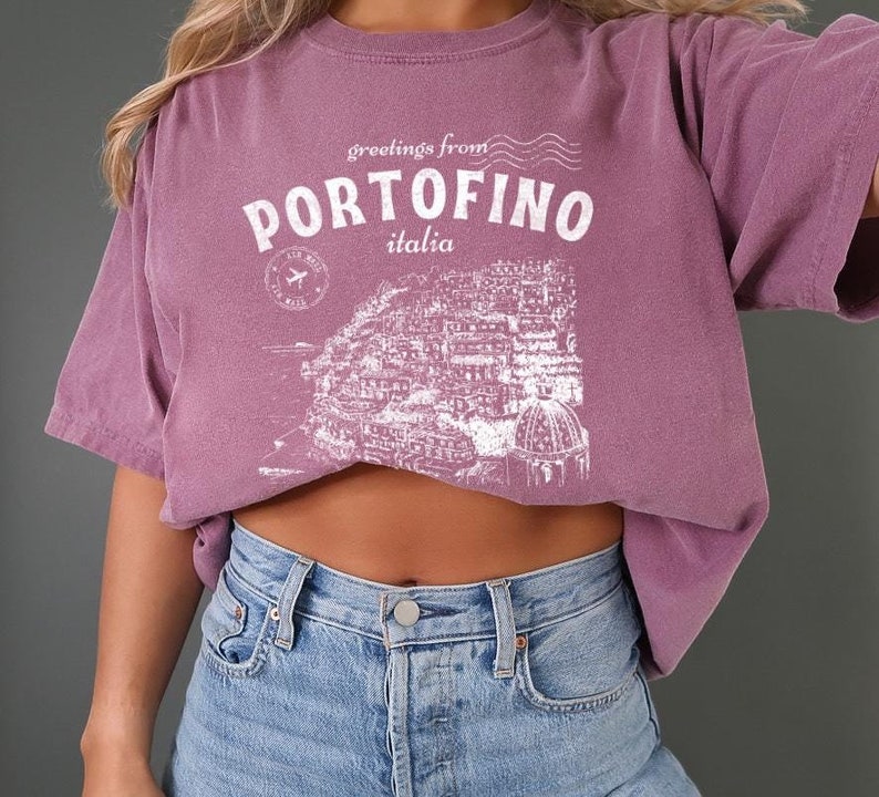 Może przedstawiać: Kr&oacute;tki, liliowy t-shirt z białym nadrukiem graficznym z tekstem "greetings from PORTOFINO italia" i stylizowaną ilustracją miasta.