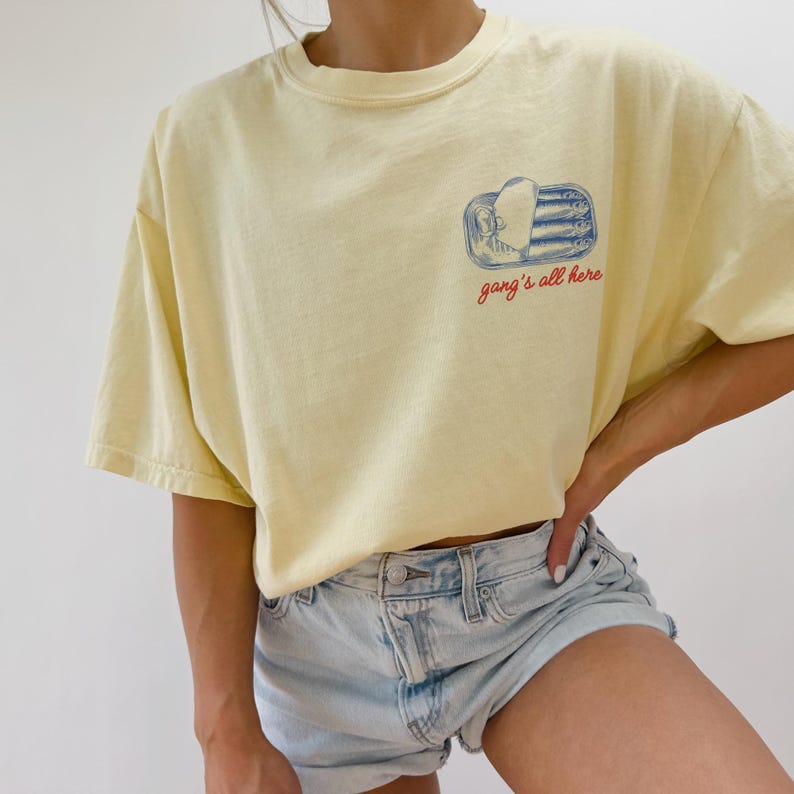 May include: A pale yellow short-sleeve t-shirt featuring a graphic on the left chest. The graphic depicts a blue tin of sardines with the text "gang's all here" written in red cursive script below the illustration of the fish inside the tin.