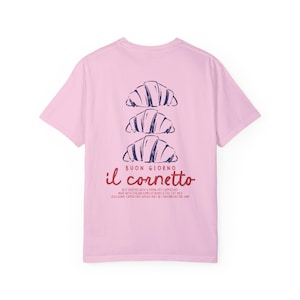 Op de afbeelding: Roze T-shirt met een afbeelding van drie croissants die op elkaar gestapeld zijn. De tekst "BUON GIORNO il cornetto" is onder de croissants gedrukt. De tekst onder de afbeelding luidt "BEST ENJOYED WITH A PIPING HOT CAPPUCCINO MADE WITH ITALIAN ESPRESSO BEANS & FULL FAT MILK.  DISCLAIMER: CAPPUCCINO SHOULD ONLY BE CONSUMED BEFORE 11AM."