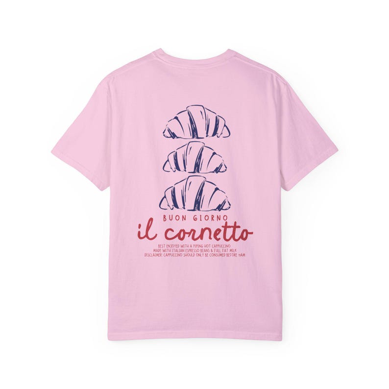 Op de afbeelding: Roze T-shirt met een afbeelding van drie croissants die op elkaar gestapeld zijn. De tekst "BUON GIORNO il cornetto" is onder de croissants gedrukt. De tekst onder de afbeelding luidt "BEST ENJOYED WITH A PIPING HOT CAPPUCCINO MADE WITH ITALIAN ESPRESSO BEANS & FULL FAT MILK.  DISCLAIMER: CAPPUCCINO SHOULD ONLY BE CONSUMED BEFORE 11AM."
