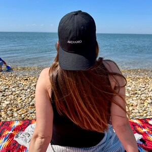 Pu&ograve; includere: Una persona indossa un cappello da baseball nero con la scritta "ANDIAMO" ricamata sul davanti, seduta su una coperta colorata sulla spiaggia.