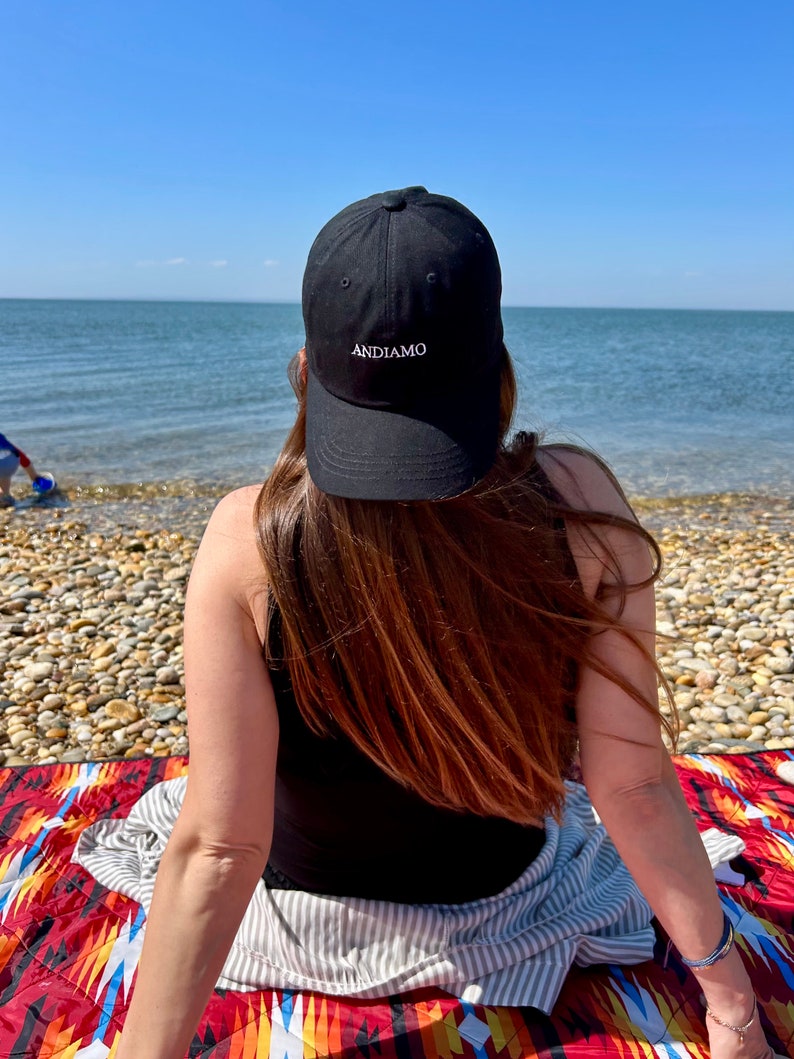 Pu&ograve; includere: Una persona indossa un cappello da baseball nero con la scritta "ANDIAMO" ricamata sul davanti, seduta su una coperta colorata sulla spiaggia.