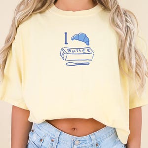 Op de afbeelding: Lichtgele T-shirt met een blauwe afbeelding met de tekst "I Butter". De afbeelding bevat een blauwe croissant, een boterpakje en een mes. Het shirt is een casual stijl met korte mouwen.
