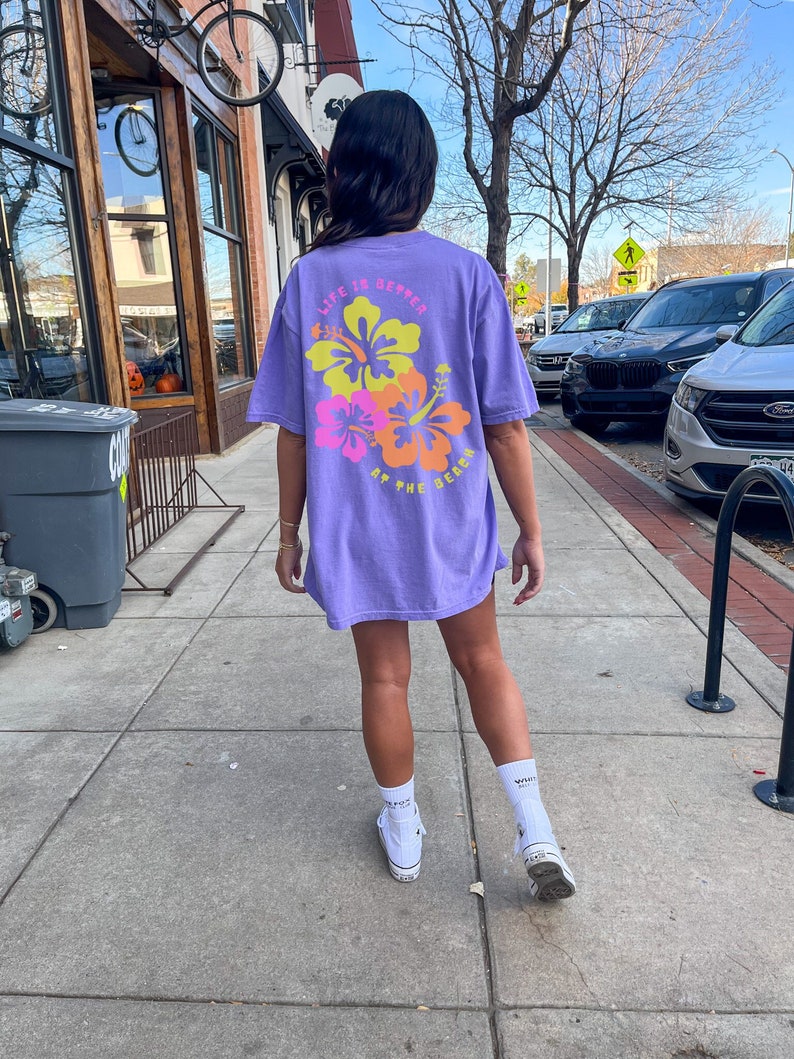 May include: A lilac-colored t-shirt with the text "LIFE IS BETTER AT THE BEACH" and a floral design in yellow, pink, and orange. The shirt is worn with white socks and sneakers.