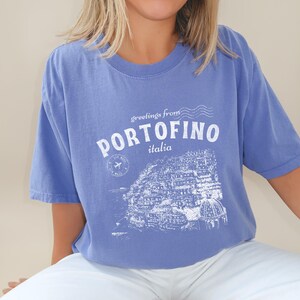 Może przedstawiać: Jasnoniebieska koszulka z białym nadrukiem graficznym przedstawiającym panoramę miasta i tekstem "greetings from PORTOFINO italia".