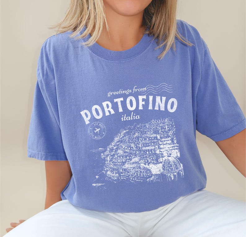 Może przedstawiać: Jasnoniebieska koszulka z białym nadrukiem graficznym przedstawiającym panoramę miasta i tekstem "greetings from PORTOFINO italia".