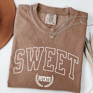 Puede incluir: Una camiseta marrón con las palabras "SWEET POTATO" en letras blancas. La camiseta tiene cuello redondo y mangas cortas. Un collar plateado con un pequeño colgante está sobre la camiseta. Un sombrero marrón y una planta seca están en el fondo.