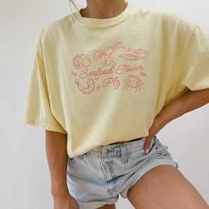 Peut inclure: Un t-shirt jaune avec un graphique rouge d'une tour de fruits de mer avec un homard, des crevettes, un crabe et du poisson. Le texte sur le t-shirt dit "Seafood Tower Plz".
