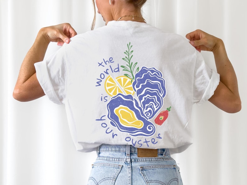 May include: White t-shirt with a graphic design of oysters, lemon, rosemary, and a red hot sauce bottle. The text "the world is your oyster" is printed on the shirt.