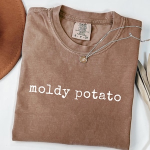 Puede incluir: Una camiseta marrón Comfort Colors con las palabras "moldy potato" impresas en blanco. Un collar plateado con un pequeño colgante está sobre la camiseta. Un sombrero marrón y una planta seca están en el fondo.