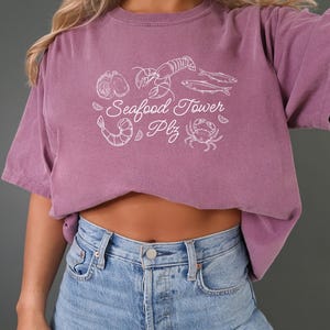 Peut inclure: Un t-shirt violet avec un motif blanc de fruits de mer, y compris un homard, des crevettes, des hu&icirc;tres, du poisson et un crabe. Le texte sur le t-shirt est "Seafood Tower Plz".