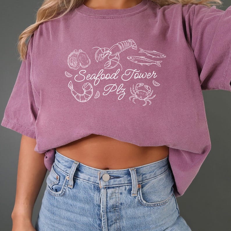 Peut inclure: Un t-shirt violet avec un motif blanc de fruits de mer, y compris un homard, des crevettes, des hu&icirc;tres, du poisson et un crabe. Le texte sur le t-shirt est "Seafood Tower Plz".