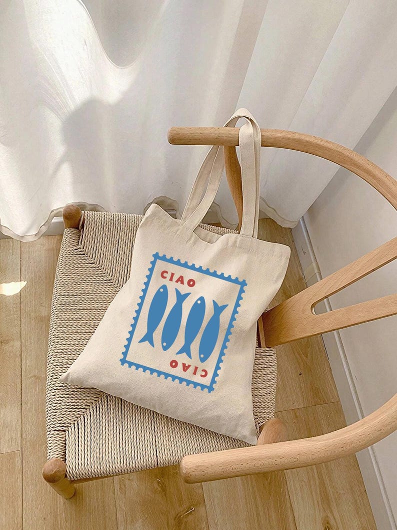 May include: A beige canvas tote bag featuring a blue and red design of three fish and the word "CIAO" above and below. The bag has two handles and is resting on a wooden chair with a woven seat.