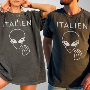 May include: Two dark gray t-shirts with the word "ITALIEN" above a white alien graphic. The word "ITALIEN" has a green, white, and red stripe above it. The shirts are worn by two people.