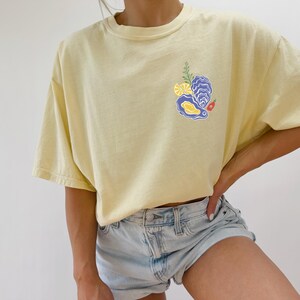 May include: A yellow short-sleeved t-shirt with a graphic of an oyster, lemon slices, and a red pepper. The shirt is worn by a person with blue denim shorts.