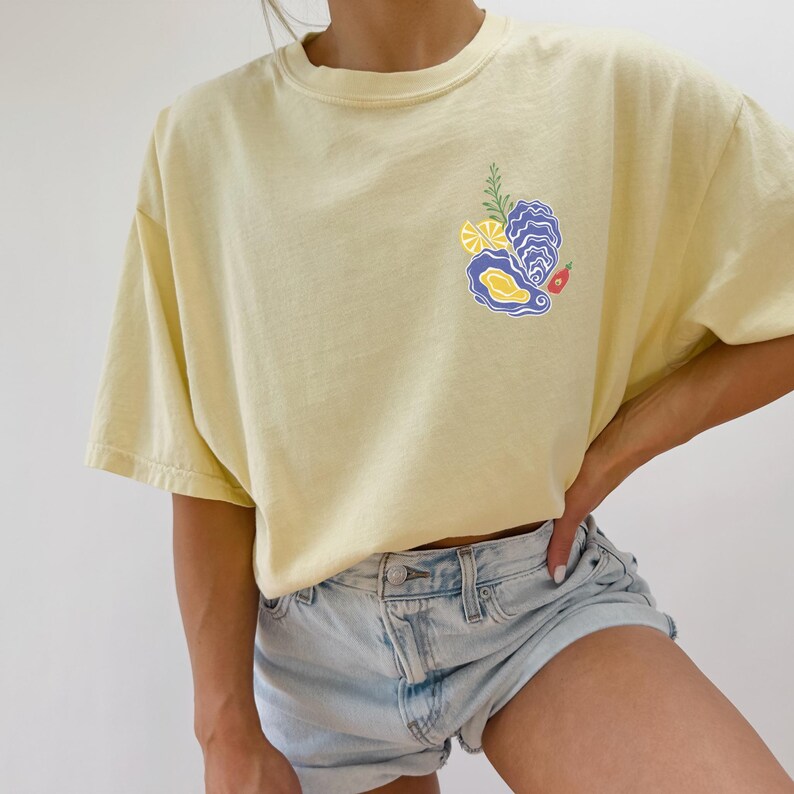 May include: A yellow short-sleeved t-shirt with a graphic of an oyster, lemon slices, and a red pepper. The shirt is worn by a person with blue denim shorts.