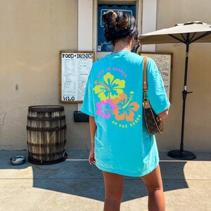 May include: A turquoise t-shirt with a floral design and the text "LIFE IS BETTER AT THE BEACH" in pink, yellow, and orange. The shirt is worn outdoors on a sunny day.