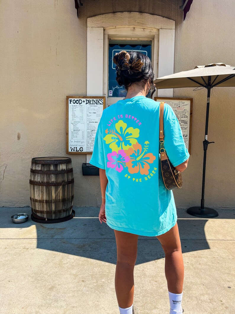 May include: A turquoise t-shirt with a floral design and the text "LIFE IS BETTER AT THE BEACH" in pink, yellow, and orange. The shirt is worn outdoors on a sunny day.