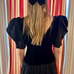 May include: A black velvet dress with short, puffed sleeves and a layered skirt. A black bow is in the person's hair. The dress has a V-neckline and a fitted bodice. The background features a striped curtain.