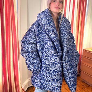 May include: An oversized, quilted puffer jacket featuring a textured, abstract pattern in shades of deep blue and light blue. The jacket has a wide, prominent collar, long sleeves, and a voluminous silhouette that extends to the upper thighs.