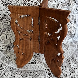 May include: A brown wooden folding stand featuring intricate carved floral and leaf patterns on two hinged panels. The structure is composed of two vertical pieces joined by metal hinges, displaying a natural wood grain and detailed relief carvings.