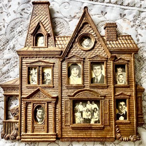 May include: A brown, house-shaped wall-mounted photo frame with a textured, wood-like finish. The frame features multiple openings for photographs, including a tall tower section, a gabled roof, and a front porch area with various portraits.