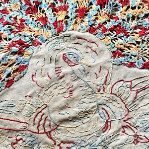 May include: A beige fabric centerpiece featuring an embroidered peacock design in red, blue, and yellow thread. The central bird is surrounded by a decorative, fan-shaped crochet border in matching red, blue, and yellow yarn, creating a circular display.