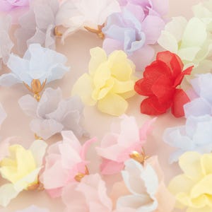 Puede incluir: Una colección de delicadas flores de tela en varios colores pastel, incluyendo azul, amarillo, rosa y rojo. Cada flor tiene un centro dorado, creando una estética encantadora y elegante. Ideal para manualidades o adornos.